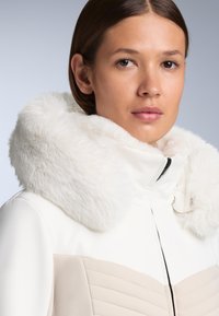 Bijela jakna s umjetnim krznom oko vrata, s naglašenim strukom, glatkom teksturom i bež panelom s_quilted detaljima. Zatvaranje na patentni zatvarač.