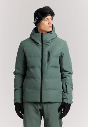 Man draagt groene gewatteerde winterjas en broek, zwarte handschoenen, zwarte muts en zwarte skibril, kijkend naar de zijkant.