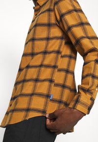 Chemise à carreaux jaunes en flanelle. Comprend des lignes bleu foncé, une fermeture à boutons et une étiquette sur le côté. Manches longues avec des poignets à bouton.