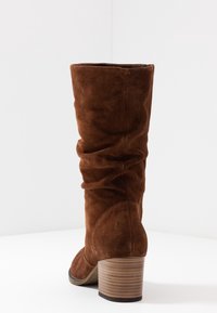 Bota de ante marrón hasta la rodilla con textura arrugada, punta redondeada y tacón de madera apilada. La parte superior es suave y ligeramente caída.