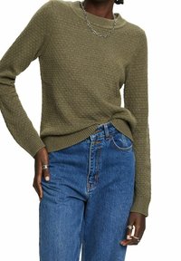 Pull en tricot texturé vert olive avec un col rond et des poignets côtelés, associé à un jean bleu taille haute. Accent de chaîne argentée au niveau du cou.