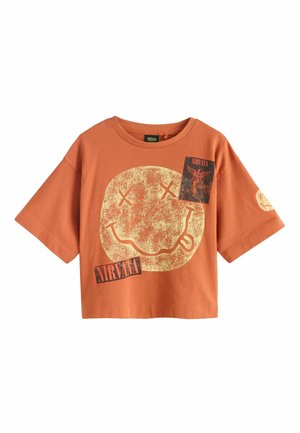 Kurz geschnittenes T-Shirt in Rostorange mit großem, abgenutztem gelbem Smiley-Gesicht und zwei rechteckigen Nirvana-Bandlogos auf der Vorderseite und dem Ärmel.