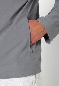 Graue Sportjacke mit strukturierter Oberfläche, seitlicher Reißverschlusstasche, enganliegenden Bündchen und entspanntem Schnitt.