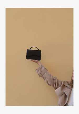 Femme en trench beige tenant un petit sac à main noir contre un mur beige uni, bras tendu vers le haut.