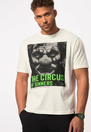 Biała bawełniana koszulka z czarną grafiką klauna i zielonym napisem "CIRCUS OF SINNERS". Noszona z czarnymi spodniami i zegarkiem z zielonym akcentem.