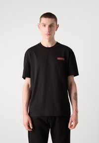 Mustasta puuvillateepaidasta, jossa on lyhyet hihat ja pyöreä pääntie. Vasemmassa rintakehässä on suorakulmainen punainen Calvin Klein -logo.