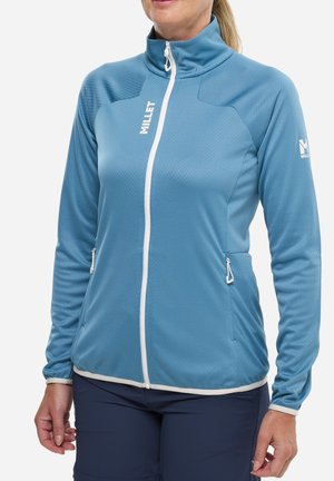 Femme portant une veste bleu clair à fermeture éclair blanche et poches, associée à un pantalon foncé, debout devant un fond uni.