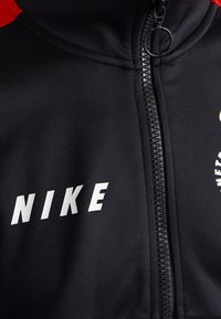 Svart zip-up-jacka med röda detaljer, med en vit "NIKE"-logotyp. Slät, stretchig tyg och en smal dragkedja med ett cirkulärt drag.