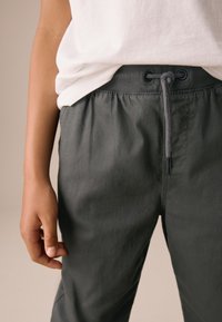 Pantalon à taille élastique gris foncé avec cordon de serrage, présentant une texture lisse et des poches latérales. Associé à un t-shirt blanc.