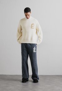 Pulover tricotat crem cu o textură în bucle și o broderie maro cu litera "E" pe piept, asortat cu pantaloni de trening gri închis cu accente albe.