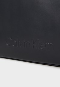 Fekete bőr kézitáska a "Calvin Klein" logóval, dombornyomott kivitelben. Letisztult dizájn, sima textúra és struktúrált forma jellemzi. Minimalista esztétika.