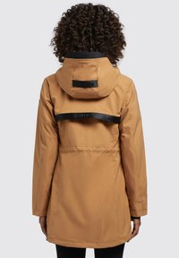 Giacca lunga con cappuccio di color beige, caratterizzata da cinturini neri in rilievo, con una texture liscia e una linea dritta. Tasche con zip sui lati.
