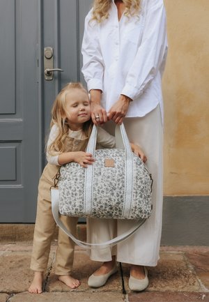 Bambino con occhi chiusi che abbraccia una borsa tipo borsone a motivo, tenuta da un adulto con abito bianco, in piedi su un pavimento di pietra vicino a una porta chiusa.