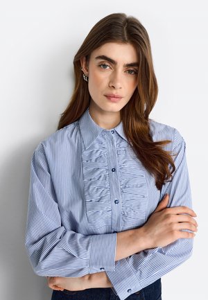 Blusa a righe blu e bianche con colletto a camicia, dettaglio plissettato sul davanti e maniche lunghe. Realizzata in tessuto leggero con una leggera lucentezza.