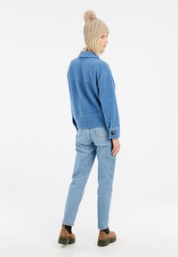 Veste en polaire bleue avec un col, associée à un jean bleu clair. Le modèle porte un bonnet en tricot beige avec un pompon. Des bottes marron complètent le look.