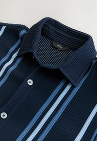 Jūros mėlynos spalvos šonkaityta marškinė su šviesiai mėlynomis ir baltomis vertikaliomis juostelėmis, su struktūruota apykakle ir dviem mygtukais. Matomas prekės ženklas.