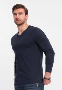 Marineblaues langärmliges V-Ausschnitt-Shirt, aus Baumwollmischung, glatte Textur, taillierte Passform, getragen mit dunklen Jeans, mit einem dezenten Handgelenksaccessoire.