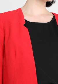 Blazer rouge à texture lisse et coupe ouverte, porté avec un haut noir. Le style sans col met en valeur la silhouette épurée.