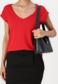 Haut rouge à manches courtes avec col en V, associé à une jupe noire ajustée. La personne tient un sac à main noir texturé avec un design géométrique.