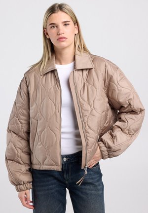 Jeune femme portant une veste matelassée beige sur une chemise blanche et un jean bleu, debout, la main dans la poche, devant un fond uni.