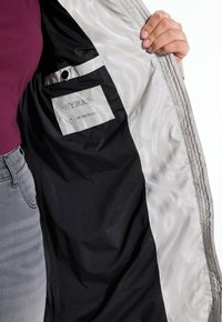 Chaqueta repelente al agua con un forro interior liso y estampado. Presenta un bolsillo negro etiquetado como "HYDRAR". El color exterior parece un beige claro.