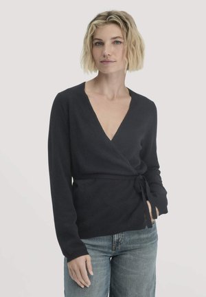 Vrouw met kort blond haar, gekleed in een zwarte wikkeltrui die op de taille is gestrikt, en blauwe spijkerbroek, staand tegen een effen lichte achtergrond.