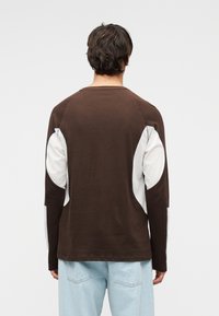 Άνδρας με μακρυμάνικο καφέ πουκάμισο με λευκά μπαλώματα στους αγκώνες και ανοιχτόχρωμο τζιν, στέκεται με την πλάτη προς το μέρος μας μπροστά σε απλό φόντο.