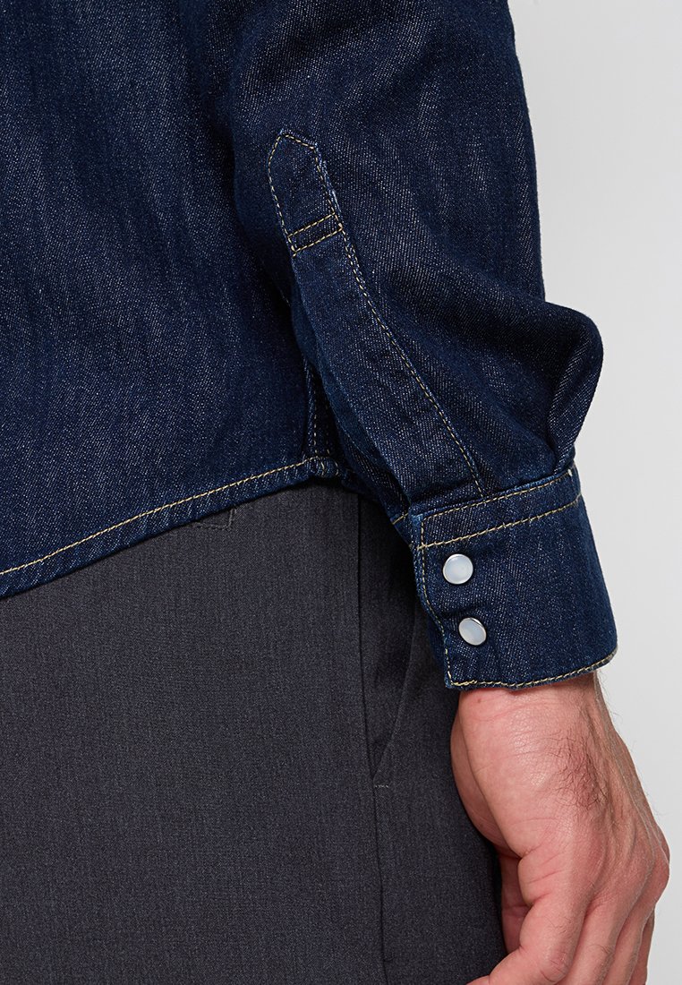 Dunkelblaue Jeanshemd mit spitzem Kragen, Druckknöpfen an den Ärmeln und kontrastierenden gelben Stichen. Glatte Textur.