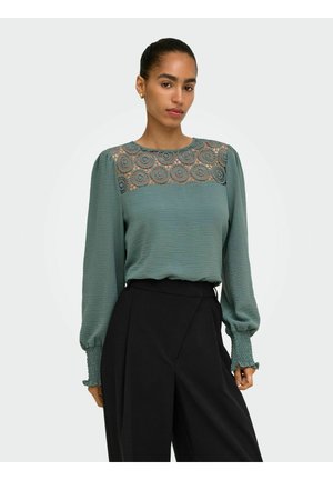 Femme portant un chemisier vert avec un détail en dentelle sur le haut de la poitrine et des manches longues bouffantes, associé à un pantalon noir taille haute.