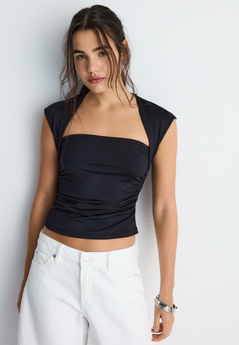 Jeune femme aux cheveux lâchés portant un haut noir moulant sans manches et un pantalon blanc, debout devant un fond clair uni.