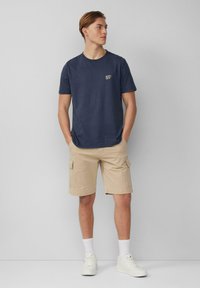 Marineblauwe katoenen t-shirt met de tekst "BEAT THE HEAT", gecombineerd met beige cargo shorts. Model draagt witte sneakers en enkelsokken.