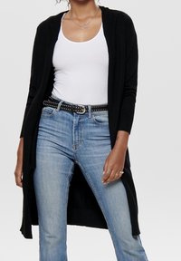 Langer schwarzer Cardigan mit geripptem Muster, offene Vorderseite, getragen über einem weißen Tanktop, kombiniert mit hellblauen Jeans und einem mit Nieten verzierten Gürtel.