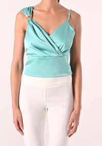 Haut en satin turquoise avec tissu drapé et accent en métal doré à l'épaule, associé à un pantalon blanc ajusté. Texture lisse, design élégant.