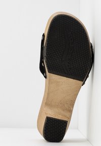 Semelle d'une sandale en bois avec une bande de roulement en caoutchouc noir, clous métalliques sur les sangles, présentée à la verticale contre un fond blanc en angle.