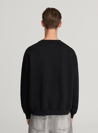 Giovane con capelli corti che indossa una felpa nera semplice e jeans di denim grigio chiaro, mostrato di spalle su uno sfondo neutro.