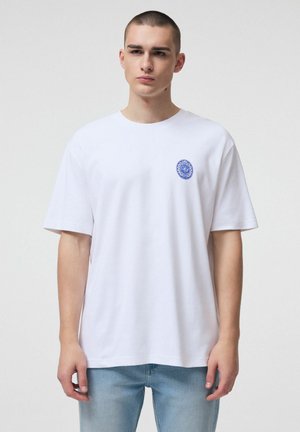 Młody mężczyzna z krótkimi włosami, ubrany w białą koszulkę z małym niebieskim okrągłym logo na lewej piersi i jasnoniebieskie dżinsy.