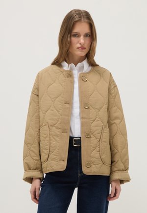 Mujer joven con una chaqueta acolchada beige, camisa blanca y vaqueros azul oscuro, de pie frente a un fondo claro y liso.