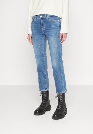 Persoon die lichtblauwe nauwsluitende jeans, een wit shirt met lange mouwen en zwarte vetercombatlaarzen draagt, staand op een witte vloer.