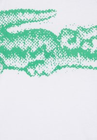 T-shirt blanc avec un motif abstrait vert ressemblant à des vagues, présentant un design texturé et moucheté dans différentes nuances de vert.