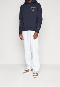 Felpa con cappuccio blu navy con logo frontale, abbinata a pantaloni bianchi larghi e sneakers multicolore con dettagli marroni, blu navy e crema.