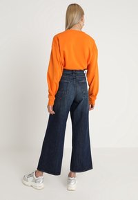 Pomarańczowa, oversize'owa bluza, wysokiej talii ciemnoniebieskie dżinsy, szerokie nogawki, białe i srebrne sneakersy, widok z tyłu.