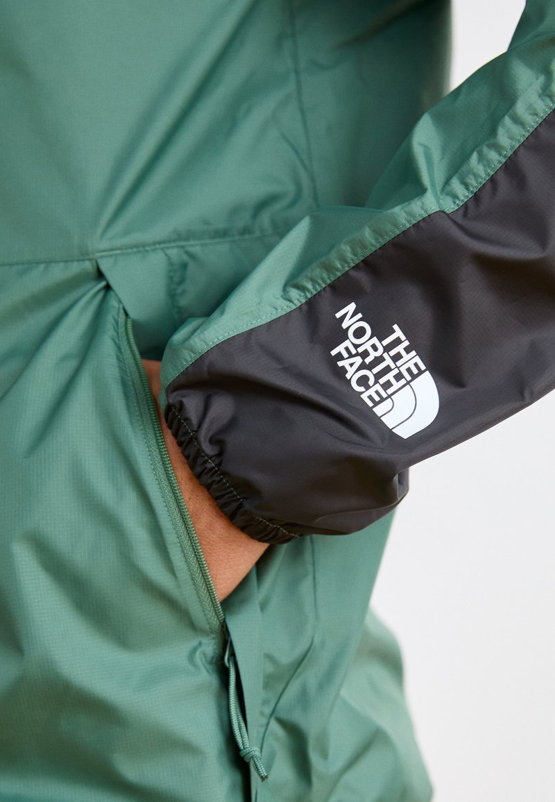 Zielona kurtka przeciwwiatrowa z czarnym elastycznym mankietem. Posiada boczny zamek błyskawiczny i logo "The North Face" w kolorze białym.