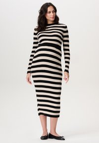 Zwangere vrouw in een zwart-beige horizontaal gestreepte midi-jurk met lange mouwen en zwarte platte schoenen, staand tegen een effen achtergrond.