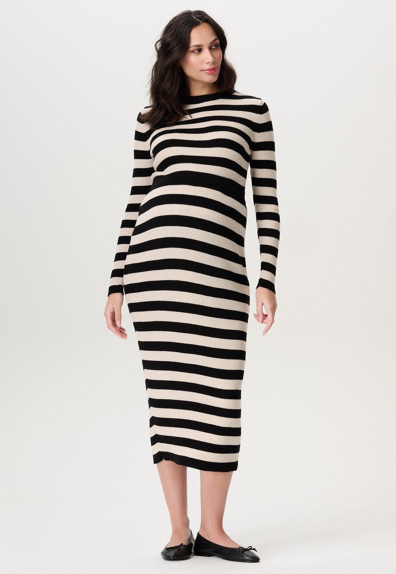 Zwangere vrouw in een zwart-beige horizontaal gestreepte midi-jurk met lange mouwen en zwarte platte schoenen, staand tegen een effen achtergrond.
