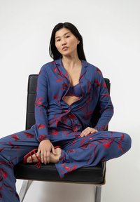 Blaues Pyjama-Set mit rotem Blumenmuster; langärmliges Hemd mit Knopfverschluss und passende Hose. Modell sitzt auf einem schwarzen Stuhl und trägt rote Sandalen.