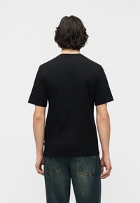 Άτομο με κοντά σγουρά σκούρα  μαλλιά που φοράει απλό μαύρο κοντομάνικο μπλουζάκι και μπλε τζιν, γυρισμένο προς τα πίσω μπροστά σε απλό φόντο.