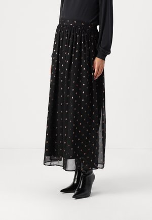 Schwarzer transparenter Maxirock mit kleinen goldenen Punkten, kombiniert mit einem schwarzen Langarmoberteil und schwarzen spitz zulaufenden Stiefeletten.