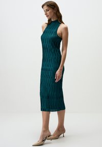 Tiefblaues ärmelloses Kleid mit vertikalem Streifenmuster, hohem Kragen und knielangem Schnitt. Glatte Textur, kombiniert mit beigen High Heels.