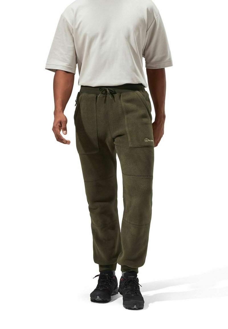 Olivgrüne Fleece-Hosen mit einem Kordelzug, seitlichen Reißverschluss-Taschen und elastischen Bündchen, kombiniert mit einem hellgrauen Kurzarmhemd und schwarzen Schuhen.