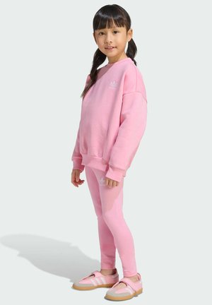 Młoda dziewczyna stoi, ubrana w różową bluzę Adidas, pasujące legginsy i różowe buty sportowe na tle jasnego, jednolitego tła.